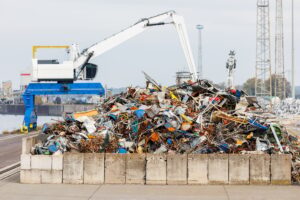 Crane at recycling facility loading large scrap metal pile with wind turbines in background.