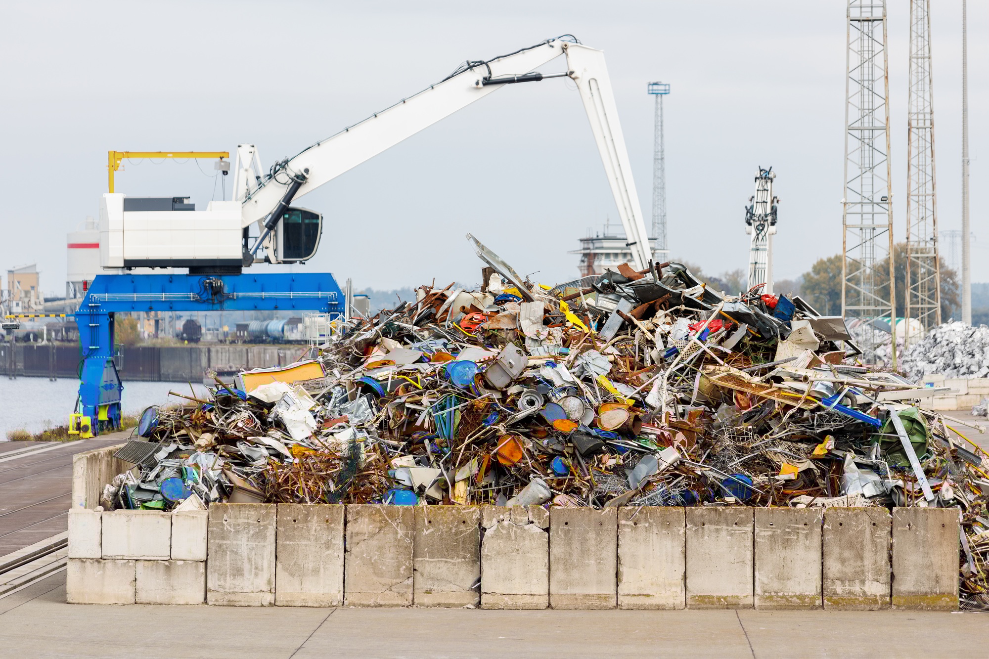 Crane at recycling facility loading large scrap metal pile with wind turbines in background.